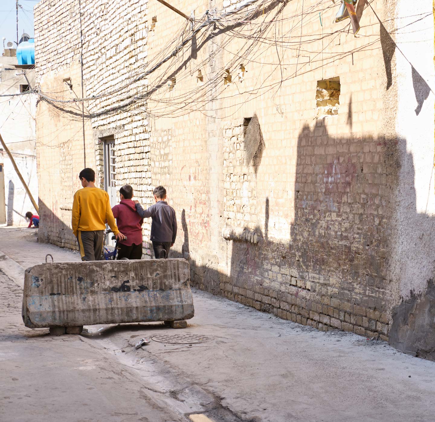 Three other children walk downstreet away from the camera, past a concrete blast barrier straddling the gutter and pavement.